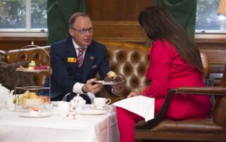 Philip Sykes sitting opposite a woman in bright red clothes and passing her a plate with fruit scones. This is a lesson in Afternoon Tea Etiquette.